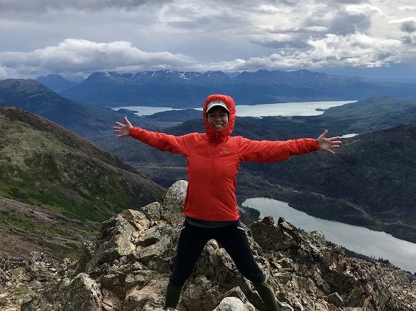 Carol Cohen standing on top of a mountain peak with beautiful lakes and rugged mountains behind her