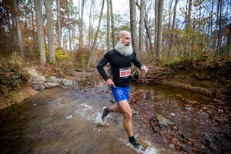 Richard Frame running through a shallow pond during a trail race. The trees in the background have fall leaves, but some of them are also bare.